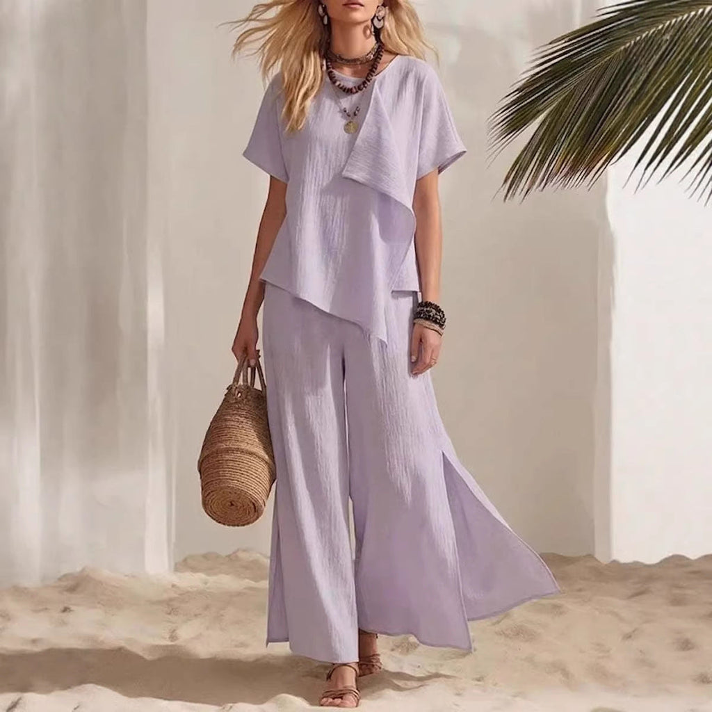 Woman in a light purple outfit standing on a sandy beach with a palm leaf in the background.